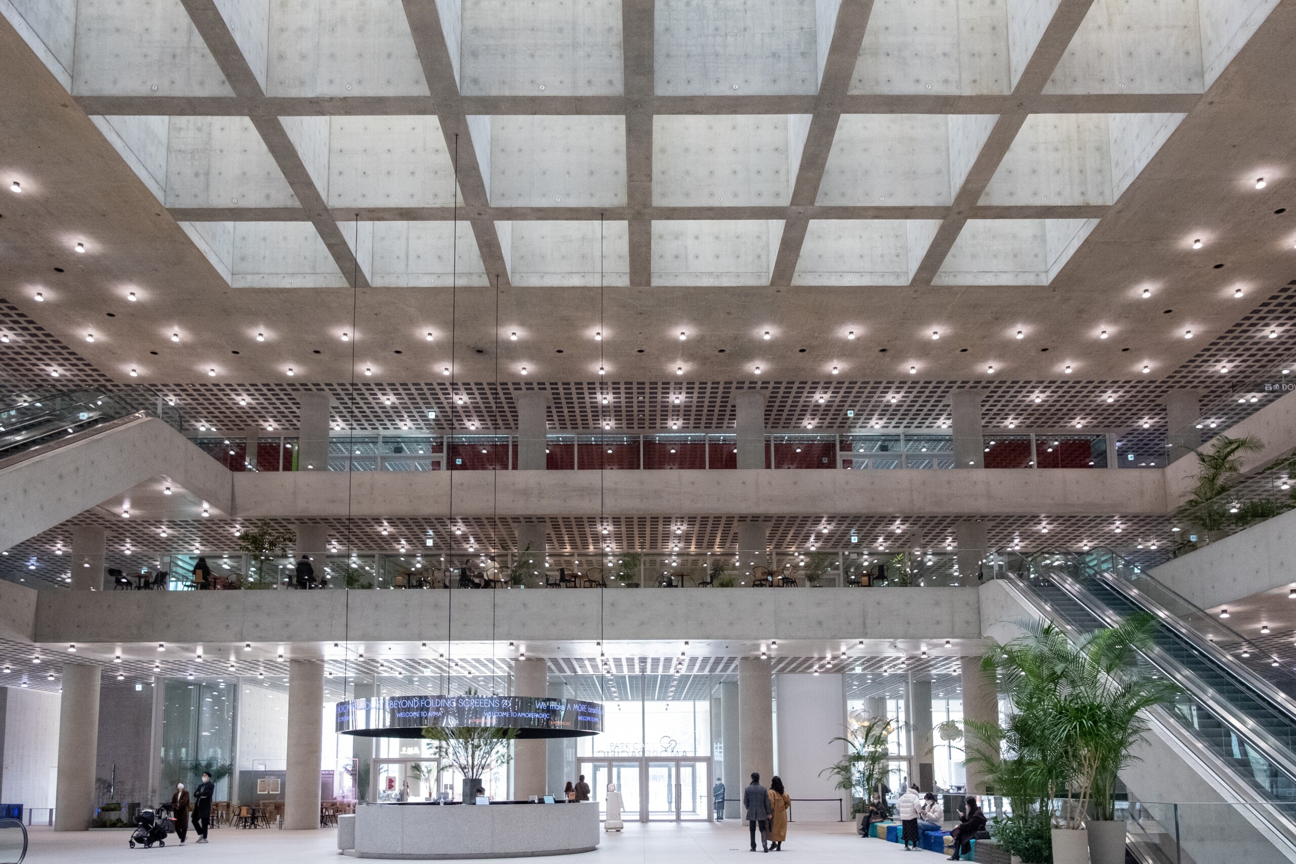 Spacious multi-storey interior of a modern public building with concrete design, large skylight ceiling, escalators, and multiple floors of open seating areas.